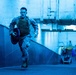 A military service member in camouflage uniform and tactical vest is running at full sprint through what appears to be an indoor corridor or tunnel, carrying a weight plate disc. The environment is bathed in a strong blue light, giving the scene a dramatic, high-intensity atmosphere. Additional personnel and equipment are visible in the background of the corridor.