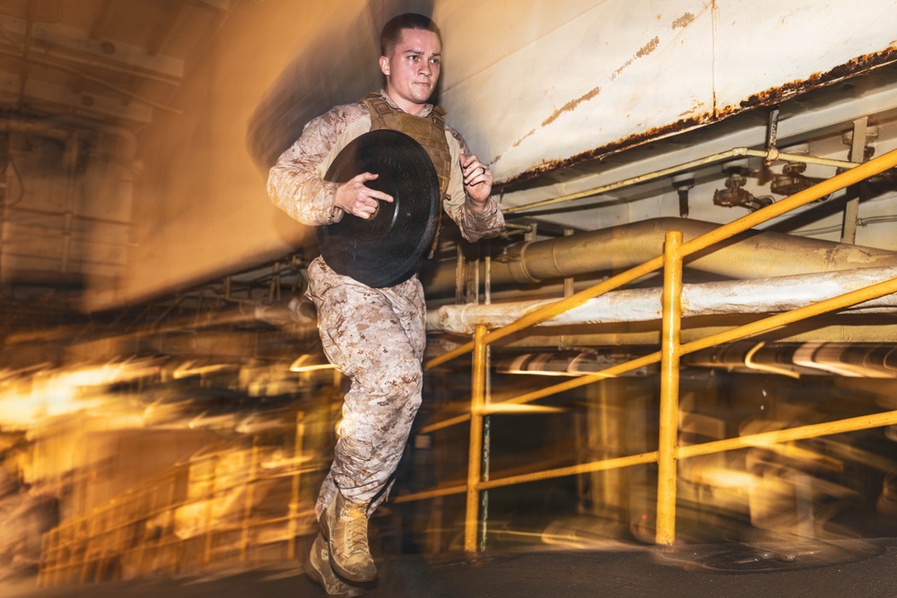 A military service member in desert camouflage (MARPAT) uniform and tactical vest is running while carrying a weight plate in an industrial or ship interior environment. The image uses motion blur and dramatic lighting to convey speed and intensity. The setting appears to be a below-deck or mechanical space with yellow safety railings and large pipes.