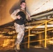A military service member in desert camouflage (MARPAT) uniform and tactical vest is running while carrying a weight plate in an industrial or ship interior environment. The image uses motion blur and dramatic lighting to convey speed and intensity. The setting appears to be a below-deck or mechanical space with yellow safety railings and large pipes.