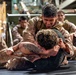 Military personnel in desert camouflage uniforms are engaged in a ground combat/grappling training exercise on a mat. One uniformed service member is on top of another who is shirtless, applying a hold while smiling. Several other uniformed personnel observe from the background in what appears to be an indoor military facility.