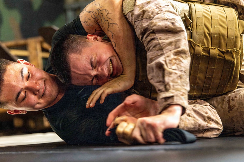 Two individuals are engaged in a ground combat training exercise. One person wearing MARPAT (Marine pattern) camouflage uniform and a tactical vest has the other person in a control hold on the ground. The person being restrained is gripping what appears to be a training knife, suggesting this is a weapons-retention or disarmament drill.