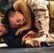 Two individuals are engaged in a ground combat training exercise. One person wearing MARPAT (Marine pattern) camouflage uniform and a tactical vest has the other person in a control hold on the ground. The person being restrained is gripping what appears to be a training knife, suggesting this is a weapons-retention or disarmament drill.
