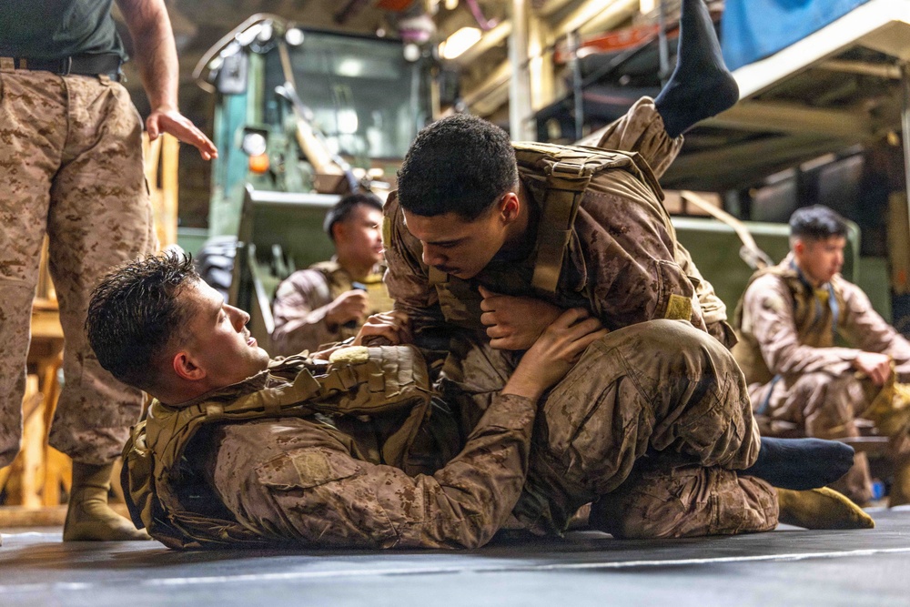 The image shows U.S. Marines engaged in close-quarters combat training, specifically ground grappling drills consistent with the Marine Corps Martial Arts Program (MCMAP). Two Marines are actively sparring on the floor in a ground control position while others observe or rest nearby. The setting appears to be the interior of a military vessel or large vehicle bay, indicated by the industrial equipment and metal flooring visible in the background.