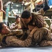 The image shows U.S. Marines engaged in close-quarters combat training, specifically ground grappling drills consistent with the Marine Corps Martial Arts Program (MCMAP). Two Marines are actively sparring on the floor in a ground control position while others observe or rest nearby. The setting appears to be the interior of a military vessel or large vehicle bay, indicated by the industrial equipment and metal flooring visible in the background.