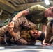 Two individuals in military attire are engaged in a ground combat training exercise on a black mat. One person wearing a green t-shirt and camouflage pants is applying a joint lock or arm control technique on another person in full camouflage uniform with a tactical vest. The setting appears to be an indoor military facility, consistent with martial arts or combatives training.