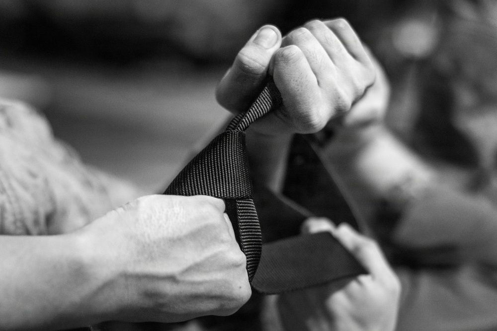 A close-up black and white photograph showing multiple hands manipulating what appears to be a nylon or webbing strap. The image is tightly cropped to focus on the hands and strap, with the background heavily blurred. The interaction suggests an adjustment or fastening action involving the textile strap.