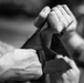 A close-up black and white photograph showing multiple hands manipulating what appears to be a nylon or webbing strap. The image is tightly cropped to focus on the hands and strap, with the background heavily blurred. The interaction suggests an adjustment or fastening action involving the textile strap.