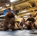 Military personnel in desert camouflage uniforms are conducting close-quarters combat or martial arts training inside what appears to be a ship's hangar bay or vehicle deck. Multiple pairs of individuals are engaged in grappling and ground fighting exercises on padded mats. The image features significant motion blur, suggesting dynamic movement during the training session.