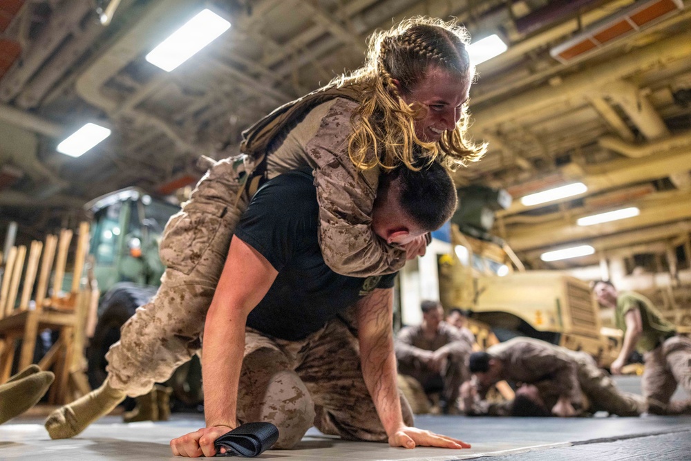 Military personnel in desert camouflage uniforms are engaged in a close-combat or martial arts training exercise in what appears to be a ship's vehicle bay or hangar deck. A female service member is on the back of a male service member who is in a crawling position, suggesting a physical fitness or combatives drill. Additional personnel can be seen in the background engaged in similar training activities.