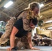 Military personnel in desert camouflage uniforms are engaged in a close-combat or martial arts training exercise in what appears to be a ship's vehicle bay or hangar deck. A female service member is on the back of a male service member who is in a crawling position, suggesting a physical fitness or combatives drill. Additional personnel can be seen in the background engaged in similar training activities.