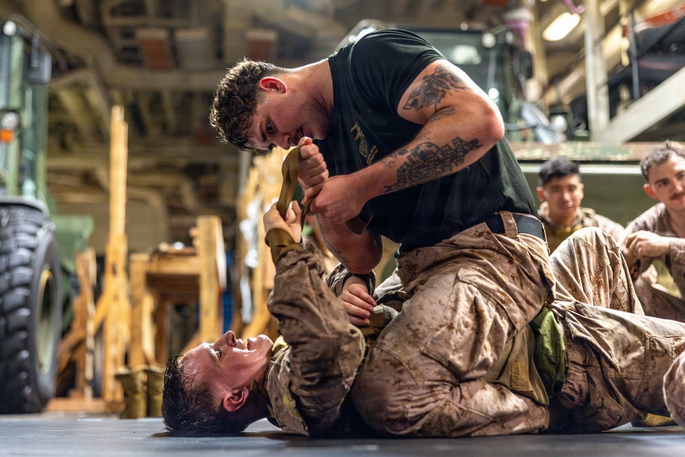 Two military personnel in combat uniforms are engaged in a martial arts or hand-to-hand combat training exercise on the floor of what appears to be a military vessel or hangar bay. One individual in a black t-shirt with visible tattoos is in a dominant ground position over another person in full desert camouflage uniform. Several other uniformed personnel observe the training session in the background.