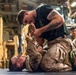 Two military personnel in combat uniforms are engaged in a martial arts or hand-to-hand combat training exercise on the floor of what appears to be a military vessel or hangar bay. One individual in a black t-shirt with visible tattoos is in a dominant ground position over another person in full desert camouflage uniform. Several other uniformed personnel observe the training session in the background.
