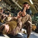 Military personnel in desert camouflage uniforms are conducting close-quarters combat or martial arts training in what appears to be a ship's vehicle deck or hangar bay. Two individuals are actively sparring on a mat, with one person on the ground and another in a dominant position applying a grappling technique. Several other service members observe the training session from the background.
