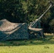 The image shows a military artillery piece partially concealed under camouflage netting in a wooded field setting. The gun barrel is elevated at a high angle, suggesting it may be positioned for use or display. The setup appears to be a field deployment with deliberate concealment measures.