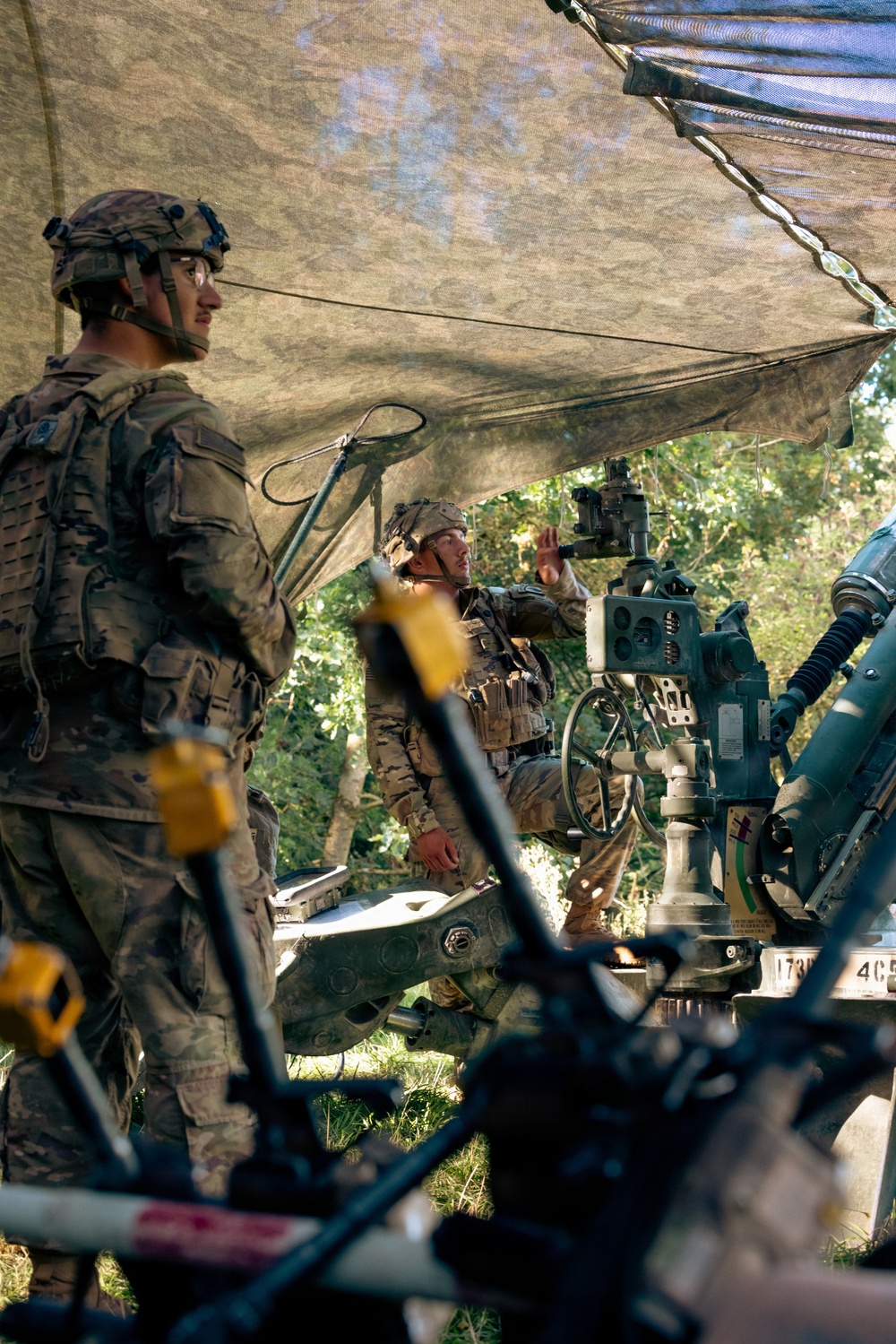 Two soldiers in modern camouflage uniforms (OCP/Multicam pattern) are operating what appears to be an M777 lightweight howitzer or similar artillery piece under a camouflage net in a wooded area. One soldier stands in the foreground wearing a combat helmet with night-vision mount, while a second soldier is seated at the gun's sighting/elevation controls. The scene suggests a field artillery training exercise or operational fire mission preparation.