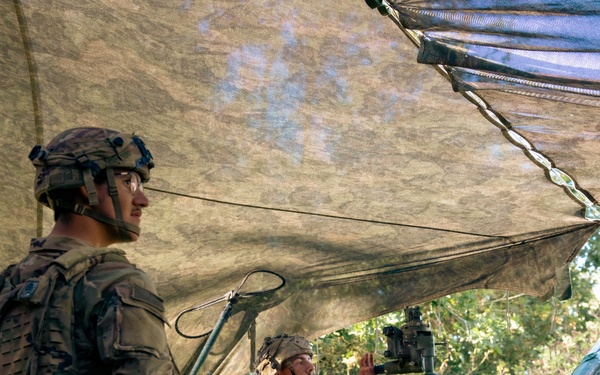 Two soldiers in modern camouflage uniforms (OCP/Multicam pattern) are operating what appears to be an M777 lightweight howitzer or similar artillery piece under a camouflage net in a wooded area. One soldier stands in the foreground wearing a combat helmet with night-vision mount, while a second soldier is seated at the gun's sighting/elevation controls. The scene suggests a field artillery training exercise or operational fire mission preparation.