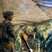 Two soldiers in modern camouflage uniforms (OCP/Multicam pattern) are operating what appears to be an M777 lightweight howitzer or similar artillery piece under a camouflage net in a wooded area. One soldier stands in the foreground wearing a combat helmet with night-vision mount, while a second soldier is seated at the gun's sighting/elevation controls. The scene suggests a field artillery training exercise or operational fire mission preparation.
