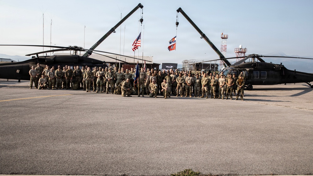 A large group of military personnel pose for a formal unit photograph on a tarmac or airfield, flanked by two UH-60 Black Hawk helicopters. Two cranes form an arch overhead, and multiple flags — including the United States flag and what appears to be a state flag — are displayed prominently in the background. The setting appears to be a military base or forward operating location.