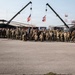 A large group of military personnel pose for a formal unit photograph on a tarmac or airfield, flanked by two UH-60 Black Hawk helicopters. Two cranes form an arch overhead, and multiple flags — including the United States flag and what appears to be a state flag — are displayed prominently in the background. The setting appears to be a military base or forward operating location.