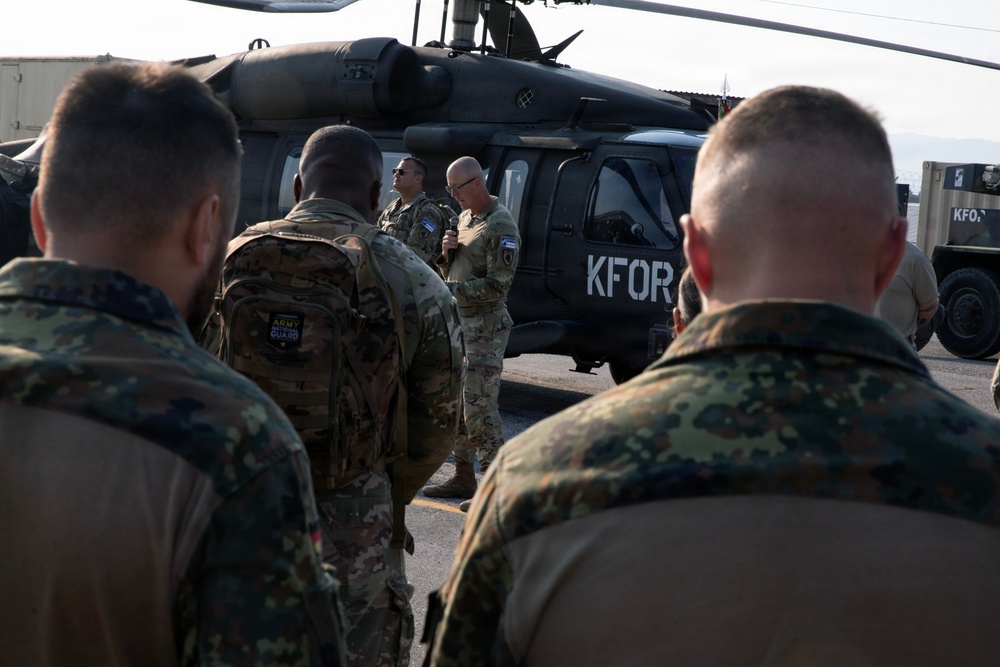 Military personnel from multiple nations are gathered in front of a KFOR (Kosovo Force) marked helicopter, likely a UH-60 Black Hawk. A soldier holding a microphone appears to be addressing the assembled group. The scene suggests a briefing or ceremony at a military airfield or base.