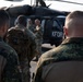 Military personnel from multiple nations are gathered in front of a KFOR (Kosovo Force) marked helicopter, likely a UH-60 Black Hawk. A soldier holding a microphone appears to be addressing the assembled group. The scene suggests a briefing or ceremony at a military airfield or base.