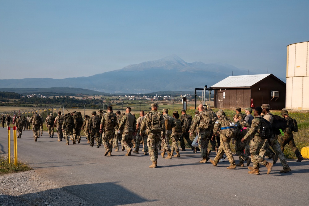 A large group of military personnel in camouflage uniforms are marching along a paved road at what appears to be a military installation or airfield. The formation moves away from the camera toward a mountainous landscape in the background. A small number of civilians appear to be integrated among the military personnel near the right side of the group.