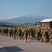 A large group of military personnel in camouflage uniforms are marching along a paved road at what appears to be a military installation or airfield. The formation moves away from the camera toward a mountainous landscape in the background. A small number of civilians appear to be integrated among the military personnel near the right side of the group.