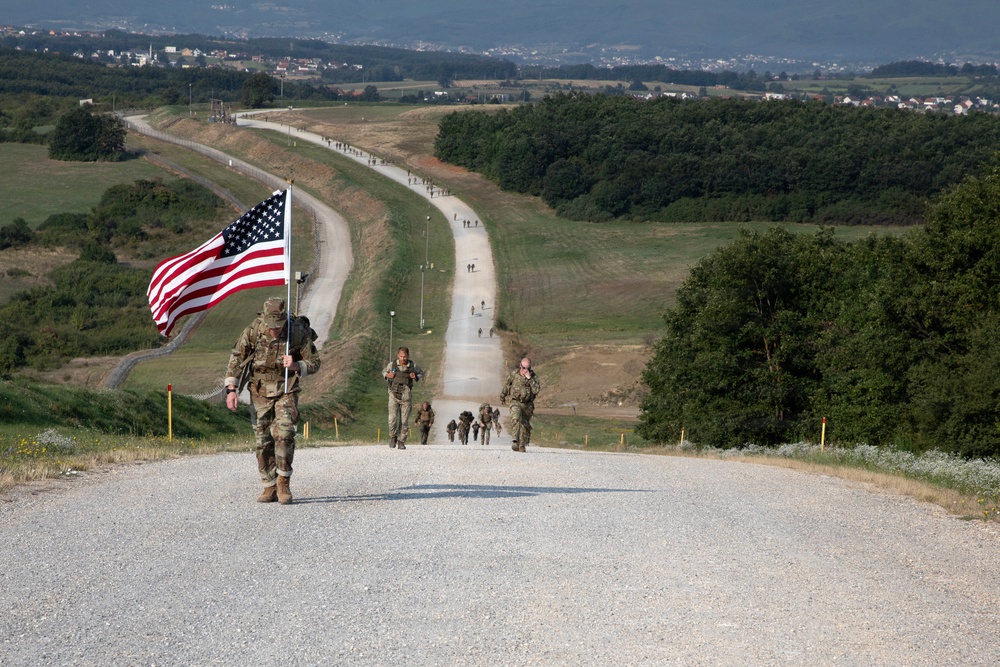 A column of military personnel in camouflage uniforms conducts a ruck march along a long straight gravel road through a rural, hilly landscape. The lead soldier prominently carries a large American flag while wearing a full rucksack. The march extends far into the distance with numerous participants visible along the road.