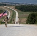 A column of military personnel in camouflage uniforms conducts a ruck march along a long straight gravel road through a rural, hilly landscape. The lead soldier prominently carries a large American flag while wearing a full rucksack. The march extends far into the distance with numerous participants visible along the road.