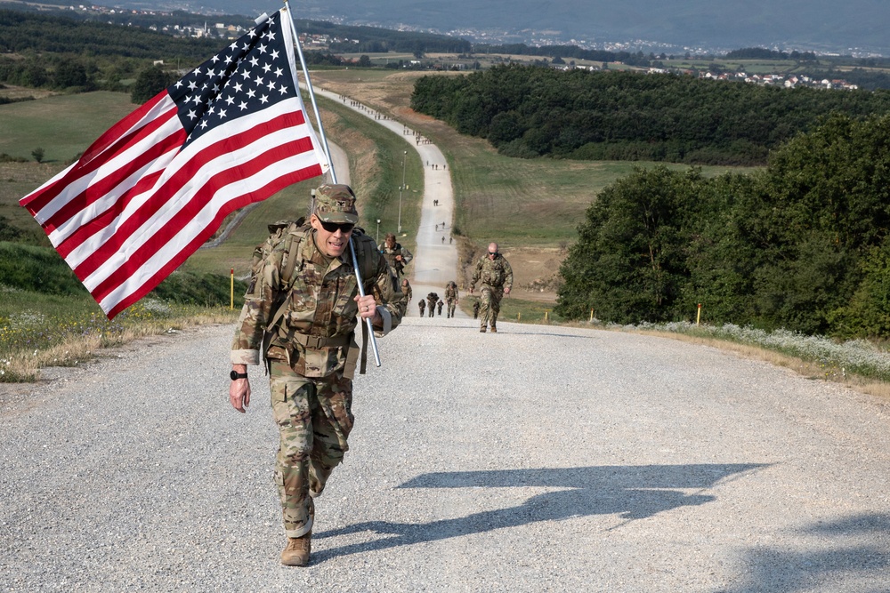 A U.S. military soldier in full combat uniform and gear carries an American flag while marching along a long straight road, with other soldiers visible behind stretching into the distance. The setting appears to be a military training area or base, surrounded by open fields and tree lines, with a town or settlement visible on the horizon. The image appears to document a ruck march or commemorative military event.
