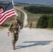 A U.S. military soldier in full combat uniform and gear carries an American flag while marching along a long straight road, with other soldiers visible behind stretching into the distance. The setting appears to be a military training area or base, surrounded by open fields and tree lines, with a town or settlement visible on the horizon. The image appears to document a ruck march or commemorative military event.