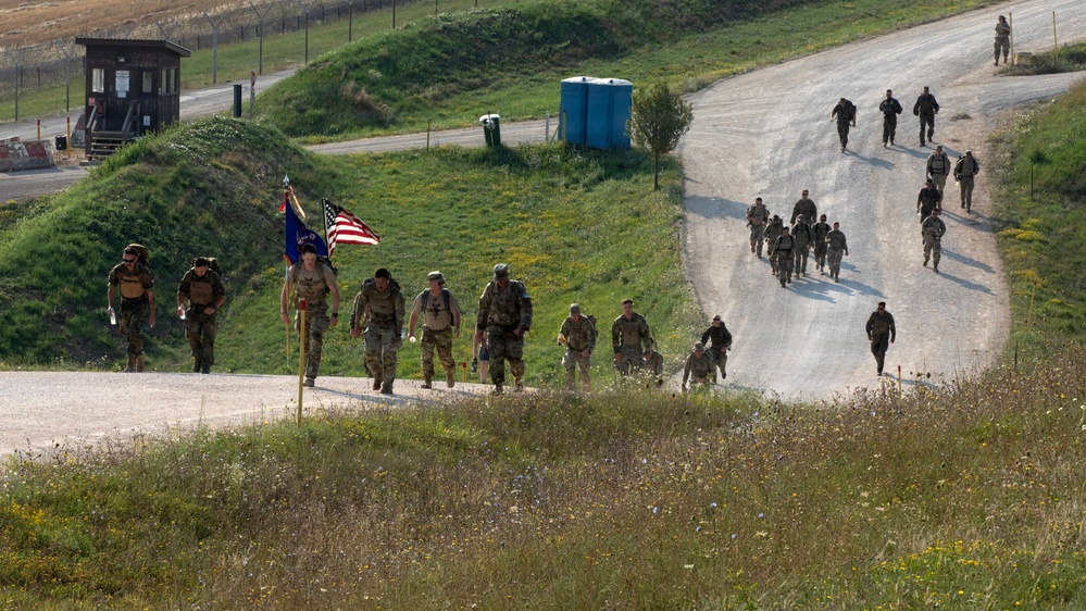 A large group of military personnel in camouflage uniforms are conducting a ruck march along a winding road at what appears to be a military installation or training base. The lead group carries an American flag and what appears to be a unit guidon, suggesting this is an organized formation march. The terrain features green hills, a guard tower, and portable facilities consistent with a military base environment.