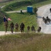 A large group of military personnel in camouflage uniforms are conducting a ruck march along a winding road at what appears to be a military installation or training base. The lead group carries an American flag and what appears to be a unit guidon, suggesting this is an organized formation march. The terrain features green hills, a guard tower, and portable facilities consistent with a military base environment.