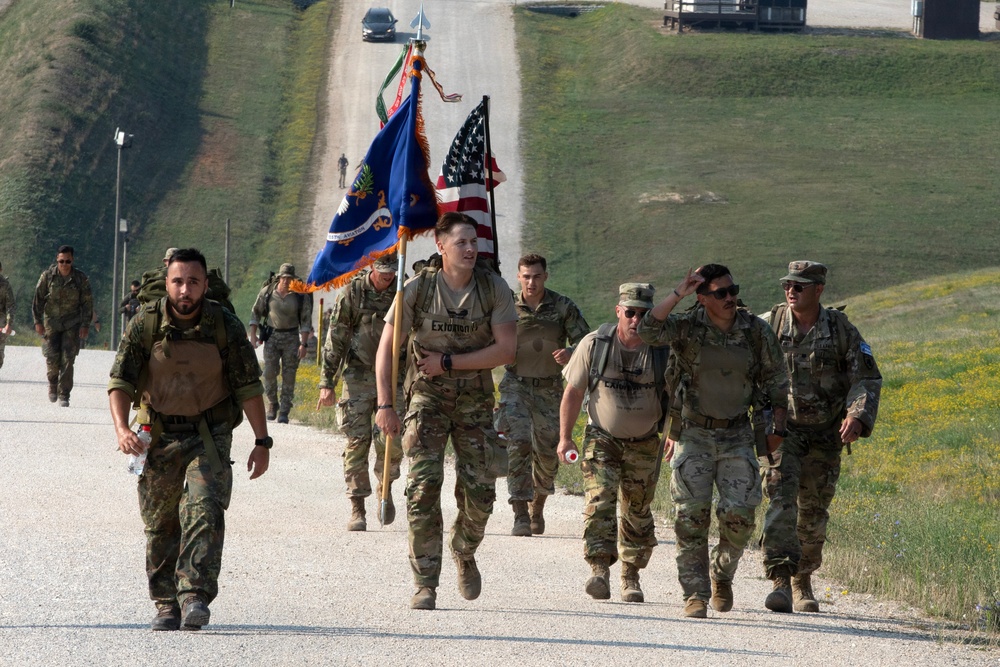 A group of military personnel in camouflage uniforms are conducting a ruck march along a paved road in a grassy, hilly terrain. One soldier in the center is carrying a blue unit guidon flag alongside a United States flag. The group appears to be participating in a formal military march or memorial event.