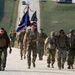A group of military personnel in camouflage uniforms are conducting a ruck march along a paved road in a grassy, hilly terrain. One soldier in the center is carrying a blue unit guidon flag alongside a United States flag. The group appears to be participating in a formal military march or memorial event.