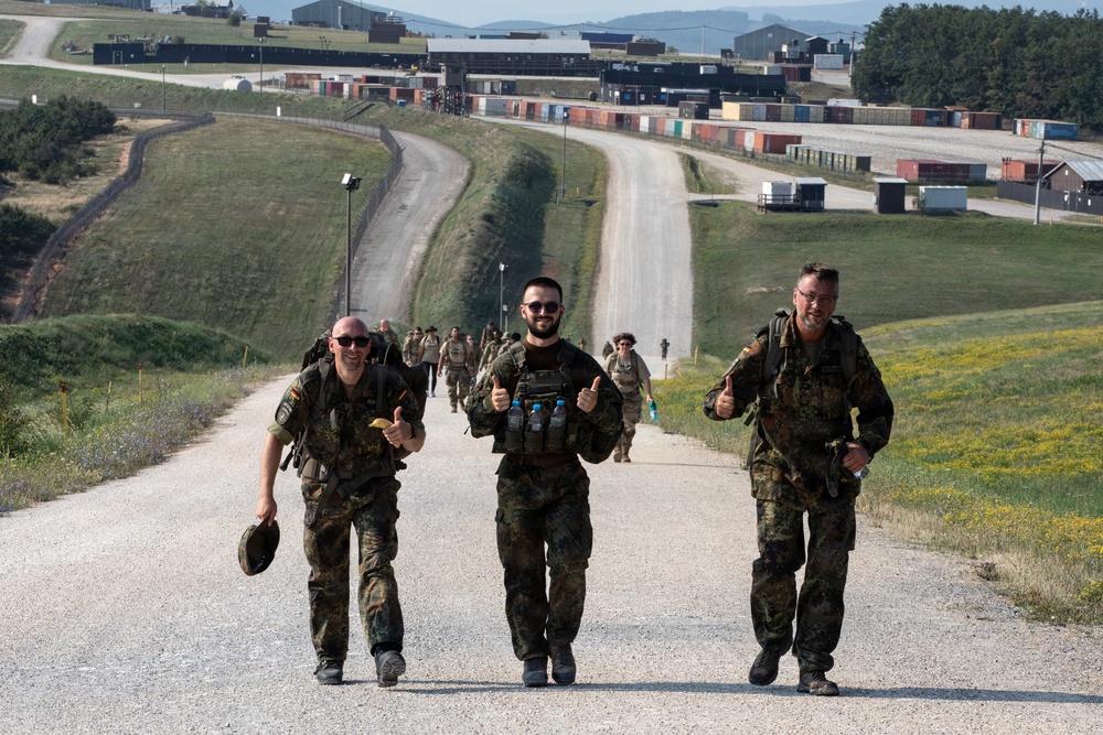 Three soldiers in the foreground march along a gravel road giving thumbs-up gestures, with a larger group of soldiers visible behind them on the same road. All personnel are wearing Flecktarn (German Bundeswehr) camouflage uniforms and carrying tactical backpacks and gear. The setting appears to be a military training area or base, identifiable by the infrastructure, shipping containers, and range facilities visible in the background.