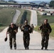 Three soldiers in the foreground march along a gravel road giving thumbs-up gestures, with a larger group of soldiers visible behind them on the same road. All personnel are wearing Flecktarn (German Bundeswehr) camouflage uniforms and carrying tactical backpacks and gear. The setting appears to be a military training area or base, identifiable by the infrastructure, shipping containers, and range facilities visible in the background.