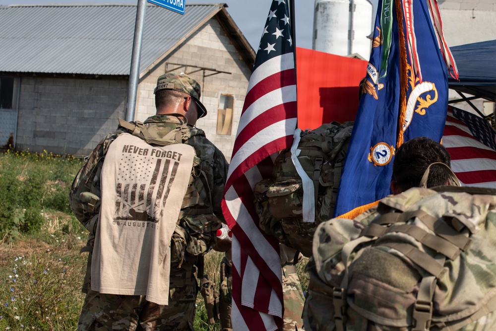 A group of military personnel in camouflage uniforms are gathered outdoors, holding the American flag and what appears to be a unit guidon or regimental flag. One soldier is wearing a 'Never Forget' commemorative t-shirt over their uniform, referencing the September 11 attacks with imagery of the Twin Towers and the date. The setting appears to be a memorial or commemorative ruck march event, likely held at a military installation or training area.