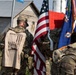 A group of military personnel in camouflage uniforms are gathered outdoors, holding the American flag and what appears to be a unit guidon or regimental flag. One soldier is wearing a 'Never Forget' commemorative t-shirt over their uniform, referencing the September 11 attacks with imagery of the Twin Towers and the date. The setting appears to be a memorial or commemorative ruck march event, likely held at a military installation or training area.