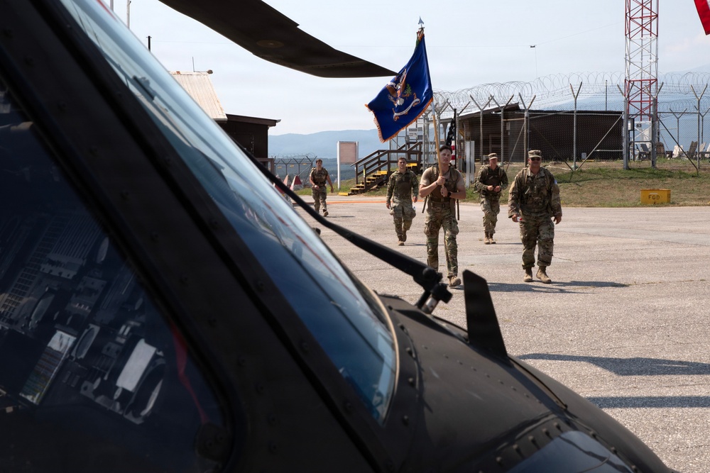 A group of military personnel in camouflage uniforms are walking across a tarmac or airfield apron toward the camera. One soldier is carrying a blue unit guidon or organizational flag. The foreground is dominated by the cockpit and rotor blade of a dark-colored military helicopter.