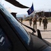 A group of military personnel in camouflage uniforms are walking across a tarmac or airfield apron toward the camera. One soldier is carrying a blue unit guidon or organizational flag. The foreground is dominated by the cockpit and rotor blade of a dark-colored military helicopter.