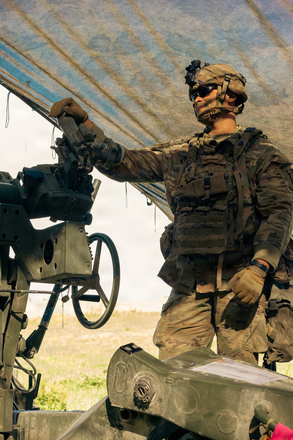 A U.S. Army soldier in full combat gear is operating or inspecting a crew-served weapon system, likely an M2 .50 caliber machine gun or similar heavy weapon mounted on a platform. The soldier is wearing Operational Camouflage Pattern (OCP) uniform, tactical vest, ballistic helmet with NVG mount, and protective eyewear. The setting appears to be an outdoor training or operational environment with a tarp providing overhead cover.