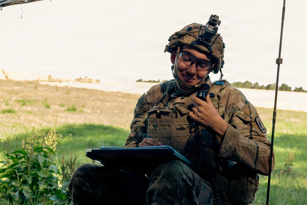 A soldier in full combat gear is seated outdoors in a field environment, simultaneously writing on a clipboard/tablet and holding a radio handset to communicate. The soldier is wearing OCP (Operational Camouflage Pattern) uniform with body armor, a tactical helmet equipped with night vision goggle mounts, and protective eyewear. The setting appears to be a field exercise or operational environment with open grassland visible in the background.