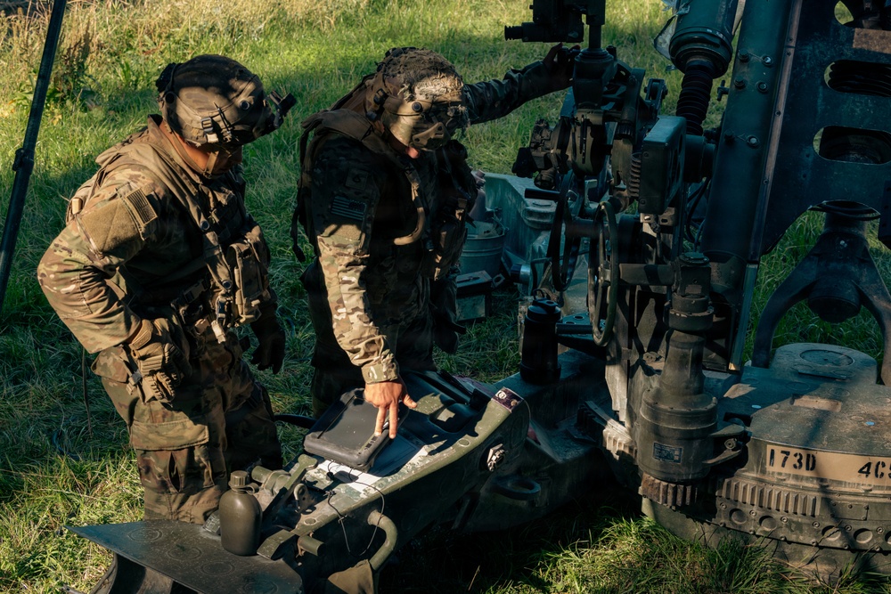 Two soldiers in full camouflage uniforms and face paint are examining equipment next to what appears to be an artillery piece in a field setting. Both personnel are wearing tactical gear including helmets with night-vision device mounts and body armor. One soldier is pointing at what appears to be a control panel or case on the ground.