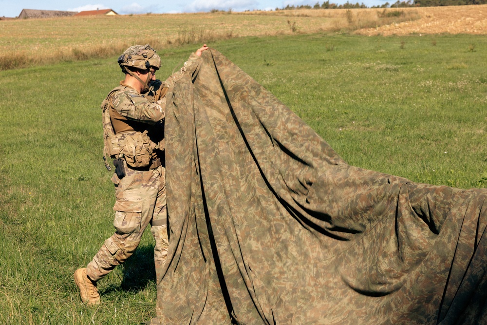 A soldier in full multicam/OCP combat uniform and tactical gear is handling a large camouflage net or tarp in an open grassy field. The soldier appears to be either deploying or folding the camouflage material, which features a woodland/green pattern distinct from the soldier's uniform. The setting appears to be a rural training area with open fields and tree lines visible in the background.
