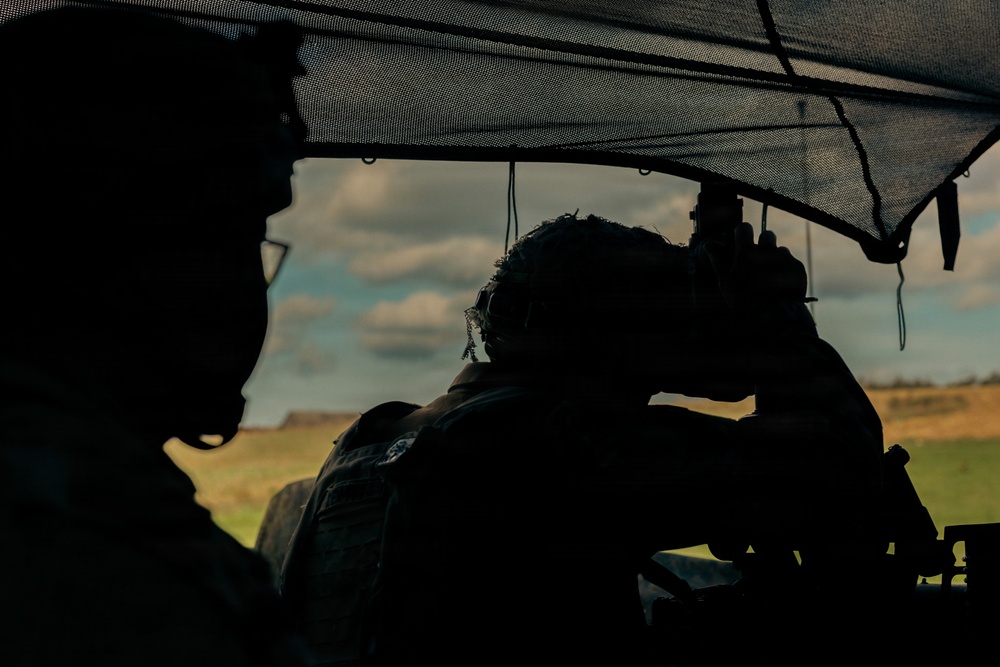 Two silhouetted figures in military/tactical gear are positioned under a mesh camouflage netting canopy. One figure appears to be looking through an optical sighting device or scope, suggesting an observation or reconnaissance activity. The background reveals an open rural landscape under a partly cloudy sky.