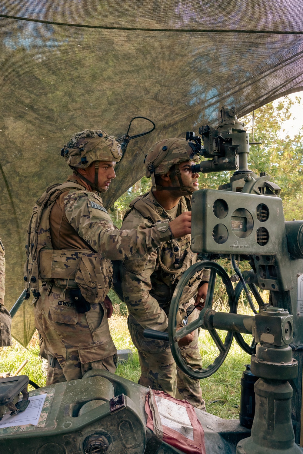 Two U.S. Army soldiers in full combat gear are operating what appears to be an artillery piece or mortar system under a camouflage net. One soldier, identified by a name tape reading 'DEWITT', is adjusting controls while the other looks through an optical sight. The setting appears to be a field training exercise in a wooded outdoor environment.