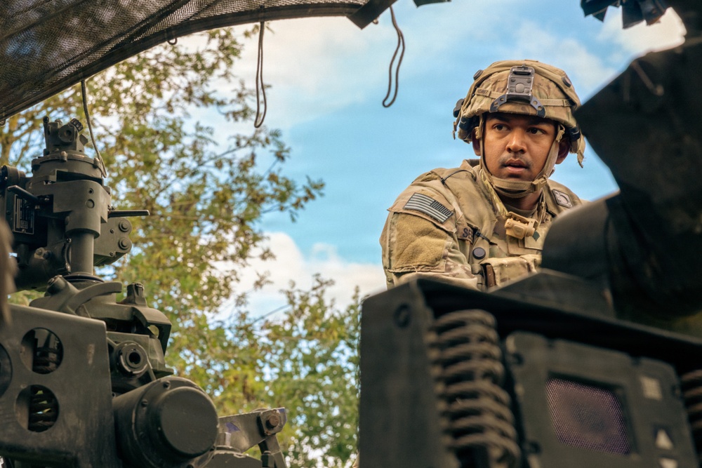A U.S. Army soldier in full combat gear is positioned inside or alongside a military vehicle, appearing alert and scanning the surroundings. The soldier is wearing MultiCam/OCP camouflage uniform with a combat helmet equipped with a night vision device mount. The low-angle photograph captures the soldier among heavy military equipment components with trees and a partly cloudy sky in the background.