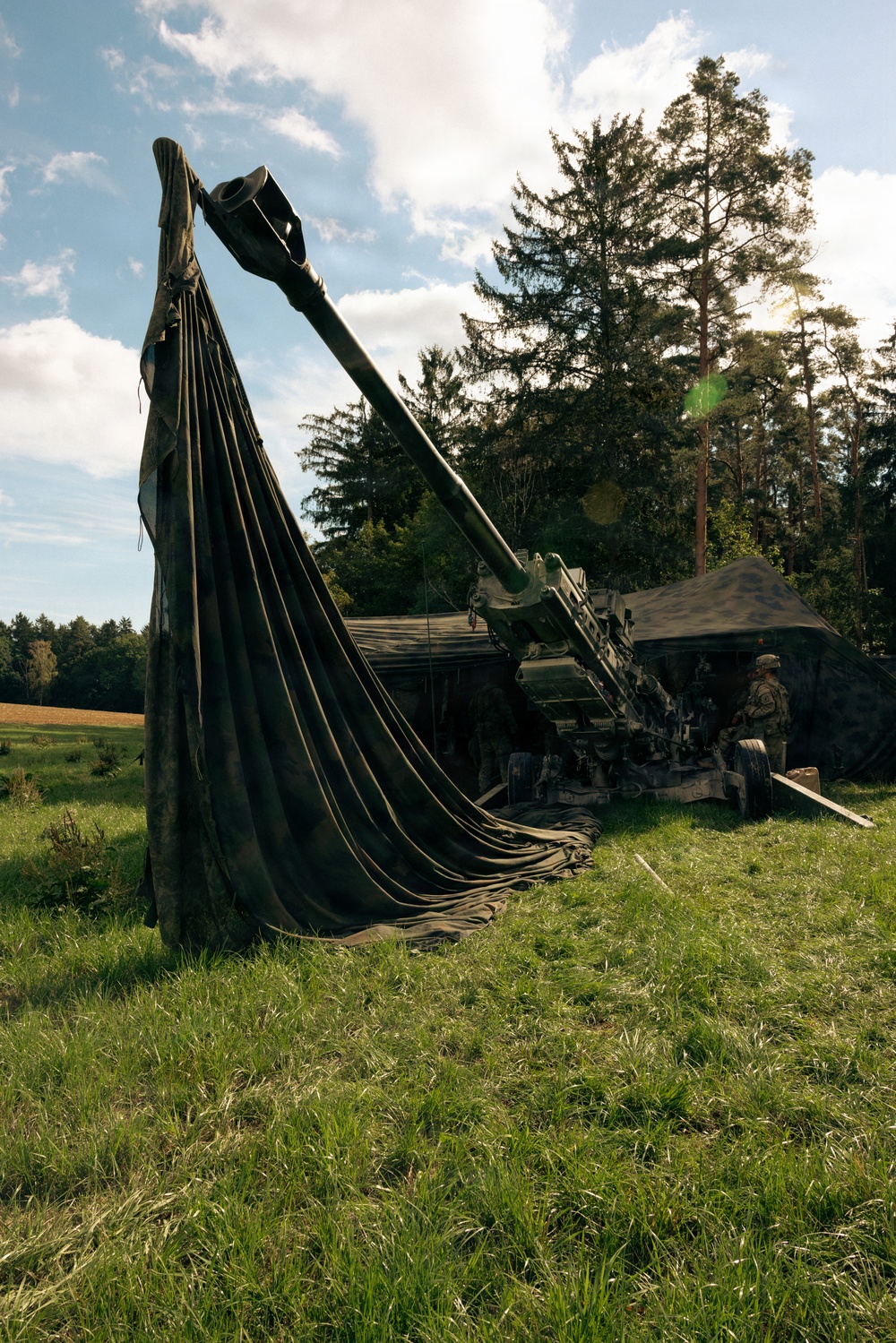 A large artillery piece, likely a howitzer, is positioned in a grassy field partially covered by a large olive-drab canvas tarpaulin draped over the barrel. At least one soldier in camouflage uniform is visible near the breech area, with a camouflage-netted shelter structure in the background. The setting appears to be a wooded, rural European landscape during daylight hours.