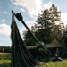 A large artillery piece, likely a howitzer, is positioned in a grassy field partially covered by a large olive-drab canvas tarpaulin draped over the barrel. At least one soldier in camouflage uniform is visible near the breech area, with a camouflage-netted shelter structure in the background. The setting appears to be a wooded, rural European landscape during daylight hours.